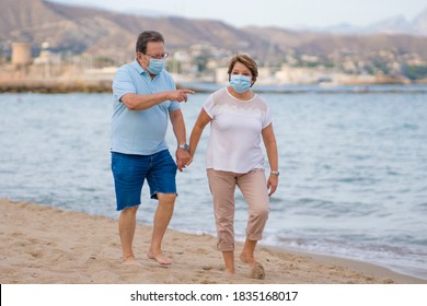 Pensioner Woman And Her Husband Taking Romantic Walk Together - Happy Retired Mature Couple In Face Mask Walking On The Beach In New Normal Holidays Trip During Covid19