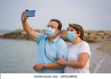 Pensioner Woman And Her Husband Taking Romantic Walk Taking Selfie - Happy Retired Mature Couple In Face Mask Walking On The Beach In New Normal Holidays Trip During Covid19