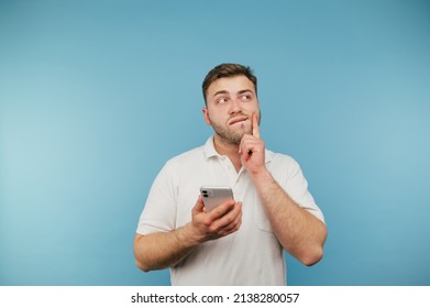 Pensive Adult Man With A Smartphone In His Hands Stands On A Blue Background And Looks Away And Thinks