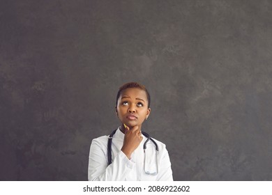 Pensive African American Female Healthcare Worker Holding Her Chin And Looking Up Thinking About Diagnosis. Serious Woman In Medical Gown On Gray Concrete Background. Medicine Concept. Place For Text.