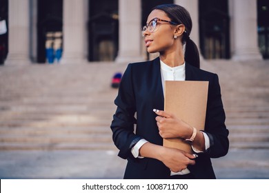 Pensive African American Female Lawyer In Stylish Formal Suit Holding Folder With Mock Up Area And Looking Away Standing Against Courthouse. Half Length Of Woman Professional Advocate With Documents