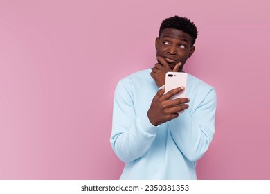 pensive african american man in blue sweater uses smartphone and plans on pink isolated background, puzzled man with phone thinks and dreams and looks at copy space - Powered by Shutterstock