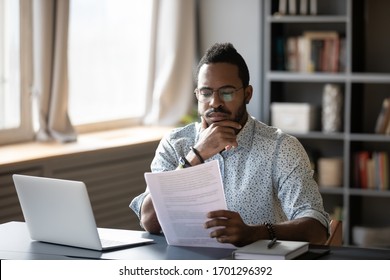 Pensive African American Man Sit At Desk Working On Laptop Reading Paper Document Thinking Or Analyzing, Thoughtful Biracial Male Worker Consider Paperwork Agreement At Office Or Home Workplace