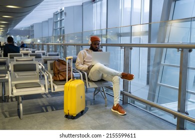 Pensive African American Man Sitting In Airport Lounge Using Laptop, Wear Headphones Listening Music. Passenger Travelling With Backpack And Suitcase, Online Working On Computer And Waiting Landing. 