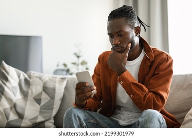 Pensive African American Man Using Smartphone, Sitting On Sofa At Home And Looking At Phone With Serious Expression, Free Space. Black Male Reading Bad News Or Browsing Internet