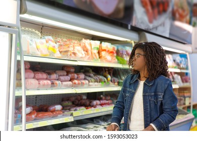 Pensive African American Woman Wheeling Shopping Cart In Grocery Store, Looking At Sausages And Ham. Buyer Shopping In Supermarket. Food Shop Concept