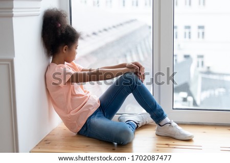 Pensive african american young girl sitting alone on windowsill in living room at home. Young introvert female person thinking looking at window. Relaxed sad melancholic daughter feel upset.