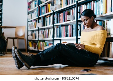 Pensive african american young woman in stylish eyeglasses for vision correction reading textbook sitting on floor near bookshelf.Concentrated dark sinned student spending leisure time in library
