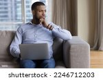Pensive African guy deep in thoughts, ponders, thinks seated on sofa with laptop staring into distance looks puzzled, uncertain distracted from tech usage, having question or challenge feels undecided