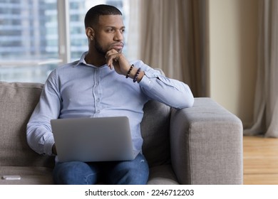 Pensive African guy deep in thoughts, ponders, thinks seated on sofa with laptop staring into distance looks puzzled, uncertain distracted from tech usage, having question or challenge feels undecided - Shutterstock ID 2246712203