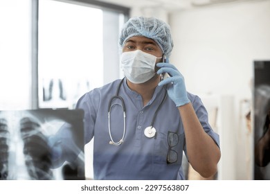 Pensive african man in face mask and scrubs holding cell phone while staying in consulting room with X-ray picture in foreground. Health professional giving recommendation about treatment via call. - Powered by Shutterstock