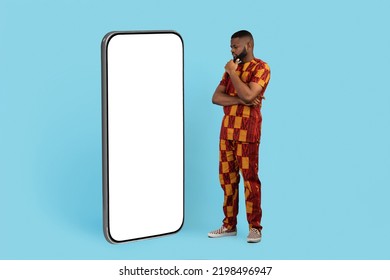 Pensive African Man In Traditional Costume Looking At Big Blank Smartphone Screen While Standing Over Blue Background, Thoughtful Black Male Thinking About Mobile Offer, Collage, Mockup