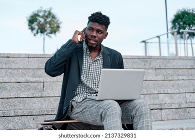 Pensive African-American Student In Suit Jacket Sitting With Laptop Open On Stone Stairs In The Street And Talking On Smartphone In Daylight.