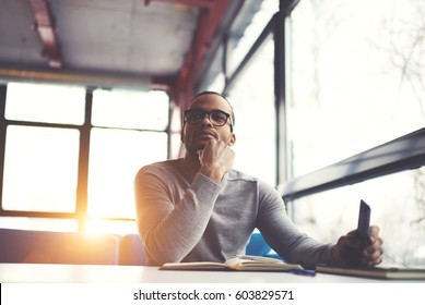 Pensive afro american handsome professional writer of popular articles in blog dressed in trendy outfit and glasses thinking over new story proofreading his script from notebook sitting in cafe