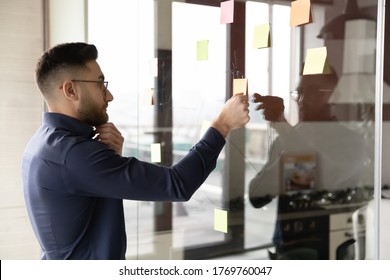 Pensive Arabic businessman brainstorm develop business plan on glass wall, work with sticky notes in office, focused Arabian male employee engaged in creative thinking in boardroom, solve problem