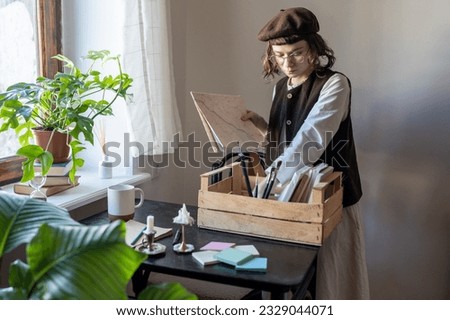 Pensive artist girl in stylish beret puts things in order in paints, creative materials, sorts out brushes, paper puts in wooden box. Art school student get ready for project. Creative hobby interest