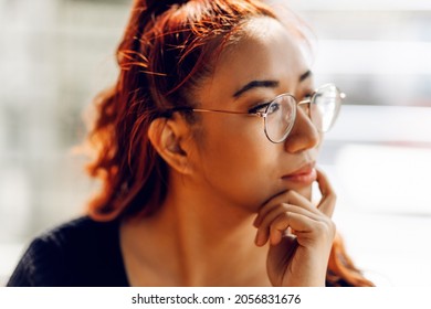 pensive Asian woman, with glasses looks out the window, with a hand on her chin, thinking about a question, obsessive expression. Smiling with thoughtful face
