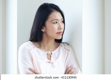 Pensive Asian woman standing at window looking out with contemplating expression. - Powered by Shutterstock