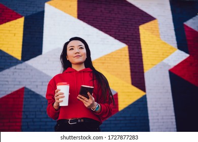 Pensive Asian youngster with takeaway cup and mobile phone thinking at urban setting with street art, trendy dressed generation Z holding cellular gadget and coffee to go spending leisure in city