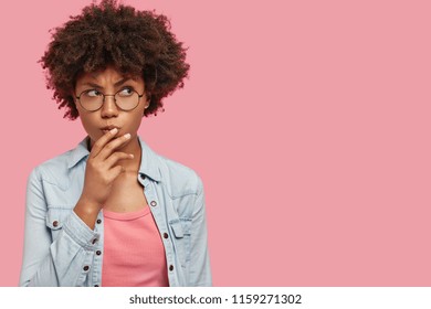 Pensive attractive curly African American female being deep in thoughts, raises eyebrows, curves lips, holds chin, wears fashionable clothes, stands against pink wall with copy space for your text