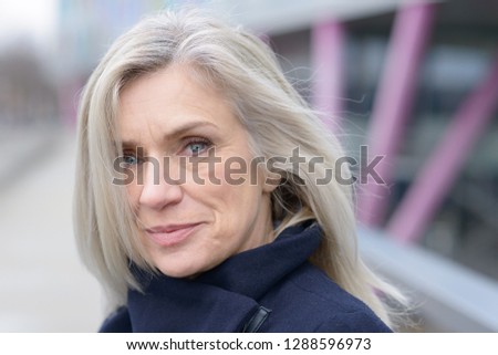 Pensive attractive woman with shoulder length blond hair scrutinising the camera with a quiet smile as she stands on an outdoor walkway in town in a close up portrait