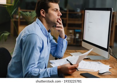 Pensive attractive young entrepreneur looking through paperwork while sitting at the desk