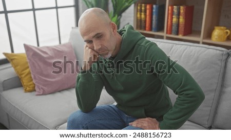 Pensive bald man with beard in green hoodie sitting on grey couch at home, reflecting alone.