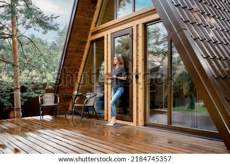 Pensive barefoot woman stands on the terrace of triangular a wooden bungalow with a glass of herbal tea