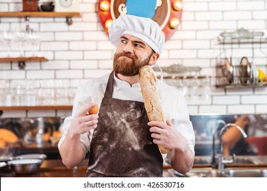 Pensive bearded baker holding eggs and bread and thinking on the kitchen