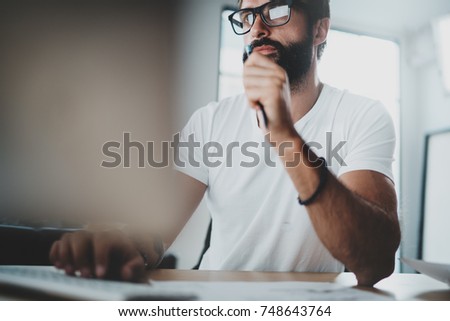 Pensive bearded coworker wearing eye glasses and working at modern loft studio-office.Blurred background. Horizontal