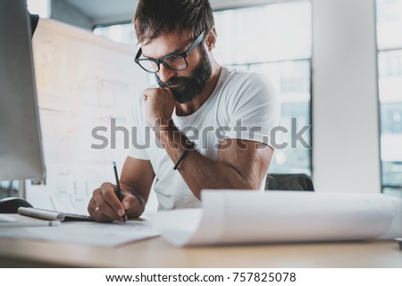 Pensive bearded designer wearing eye glasses and white tshirt, working at modern loft studio-office.Man drawing scetches.Blurred background. Horizontal