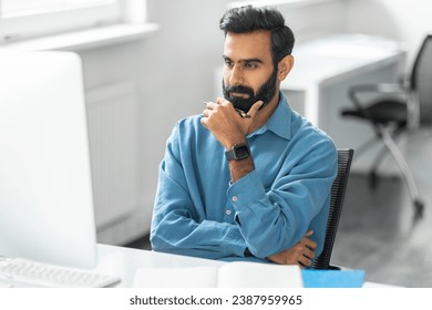 Pensive bearded indian man wearing blue shirt and smartwatch, touching chin and looks intently at computer screen in modern, bright office setting - Powered by Shutterstock