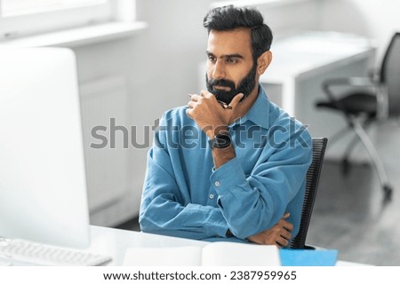 Pensive bearded indian man wearing blue shirt and smartwatch, touching chin and looks intently at computer screen in modern, bright office setting