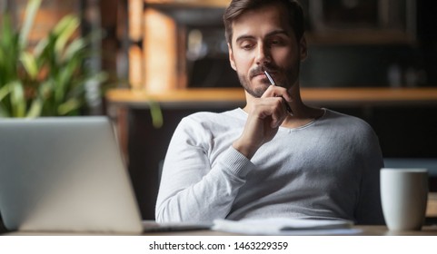 Задумчивый бородатый мужчина, сидящий за столом, пьет кофе, работает за ноутбуком, думает о решении проблемы, вдумчивый сотрудник-мужчина размышляет над идеей, глядя на решение на экране компьютера