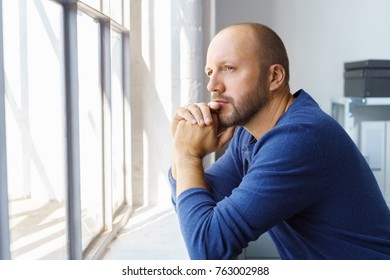 Pensive bearded man standing watching through a window resting his chin on his hands with a speculative expression - Powered by Shutterstock