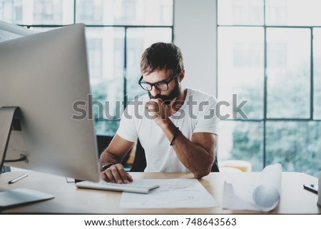 Pensive bearded man wearing eye glasses and working at modern loft studio-office.Blurred background. Horizontal