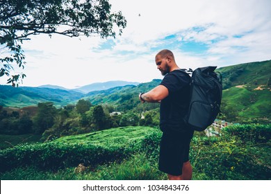 Pensive bearded tourist with backpack looking at smartwatch to tracking GPS walking in green locality of asia mountains.Hiker with rucksack managing tame on wristwatch during adventure trek