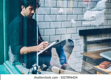 Pensive Bearded Young Man Dressed In Black Shirt Writing Down Essay In Notepad Resting In Coffee Shop Interior.Skilled Hipster Guy Writer Noting Some Records In Notebook Sitting In Coworking Space