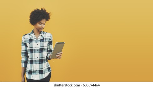 Pensive beautiful black teenage girl in checkered shirt with curly afro hair on yellow empty background holding digital tablet with copy space for text, your logo or advertising