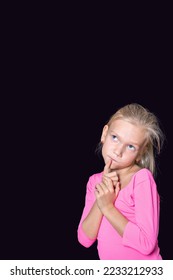 Pensive beautiful caucasian girl in a pink sweater looking up and holding her finger to her face on a black background, copy space. A template for overlaying your text. - Shutterstock ID 2233212933