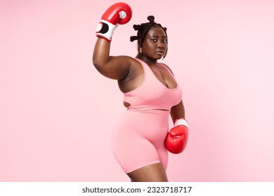 Pensive beautiful Nigerian woman wearing sports uniform red boxing gloves looking at camera isolated on pink background. Authentic body positive African model posing for picture, copy space - Powered by Shutterstock