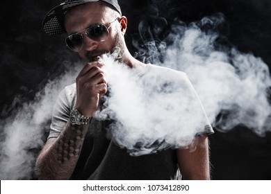 Pensive Black bearded male dressed in a grey shirt, sunglasses and baseball cap vaping. man in holding a mod. A cloud of vapor. Black background.
