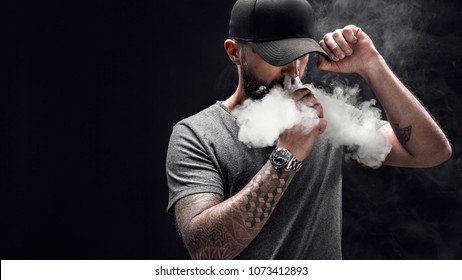 Pensive Black bearded male dressed in a grey shirt, sunglasses and baseball cap vaping. man in holding a mod. A cloud of vapor. Black background.