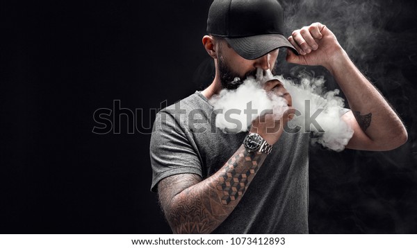 Pensive Black bearded male dressed in a grey\
shirt, sunglasses and baseball cap vaping. man in holding a mod. A\
cloud of vapor. Black\
background.