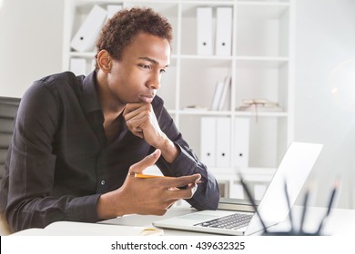 Pensive black businessman working on project on office desk with laptop and notepad. Bookshelf with documents in the background