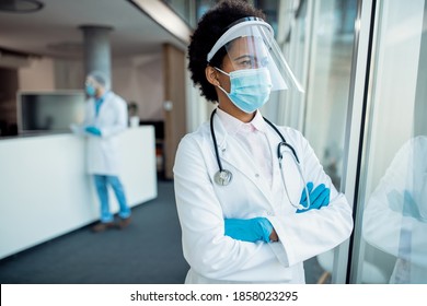 Pensive Black Female Doctor Looking Through The Window While Wearing Face Shield And Protective Mask During COVID-19 Pandemic At Medical Clinic. 