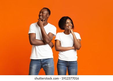 Pensive Black Man And Woman Standing Next To Each Other And Thinking, Touching Their Chins And Smiling, Copy Space. Young African-american Couple Day Dreaming Together On Orange Studio Background