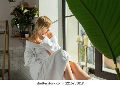 A Pensive Blonde Girl In A White Robe And With A Towel On Her Head In The Bathroom Sits On The Windowsill, A Young Relaxed Woman In The Bathroom Touches Her Hair, A Sunny Summer Day At The Window 