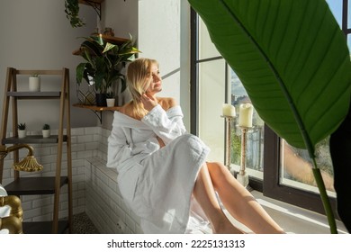 A Pensive Blonde Girl In A White Robe And With A Towel On Her Head In The Bathroom Sits On The Windowsill, A Young Relaxed Woman In The Bathroom Touches Her Hair, A Sunny Summer Day At The Window 