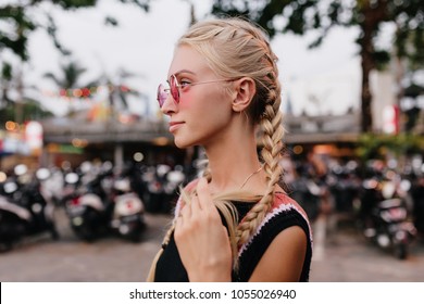 Pensive blonde woman in black attire posing on blur street background. Outdoor shot of serious tanned lady with braids wears pink sunglasses.